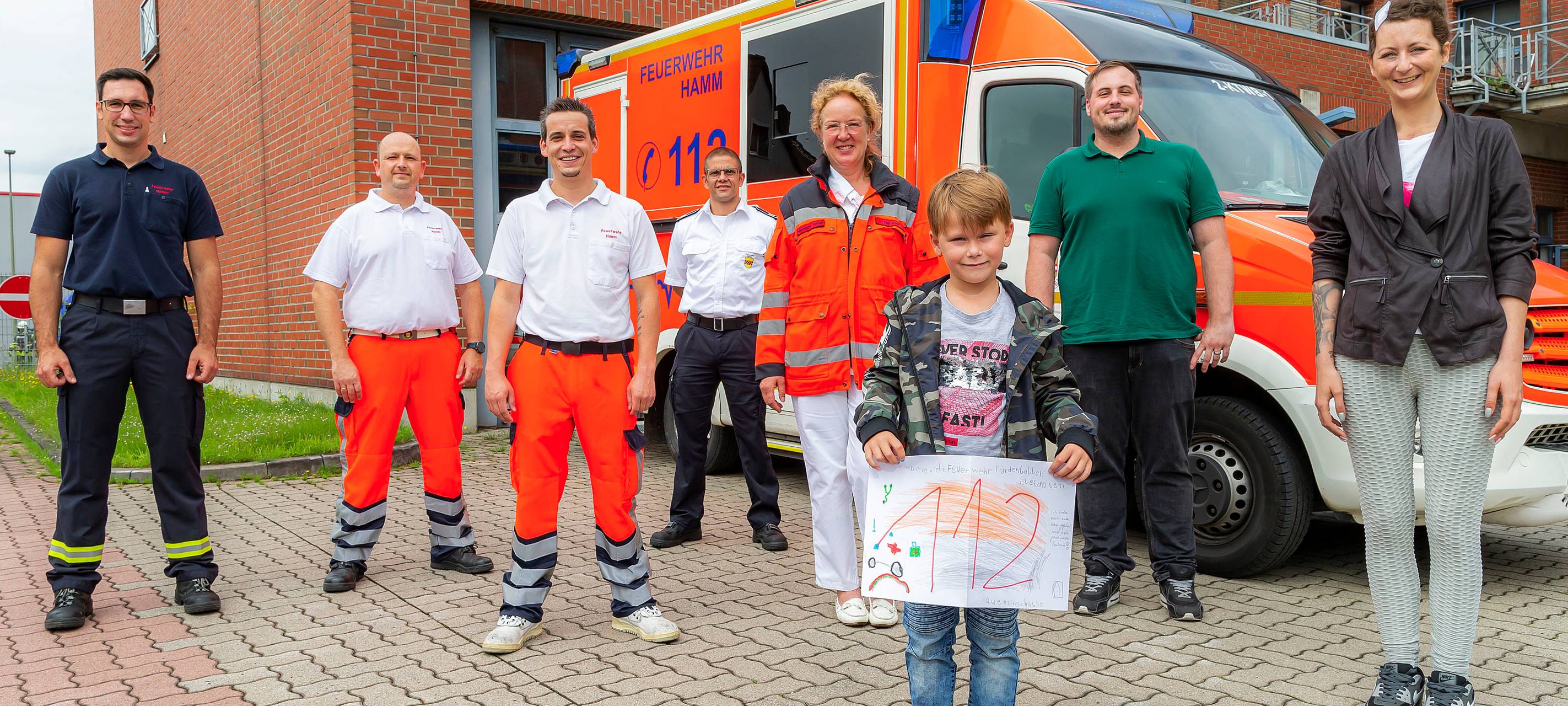 Quentin steht mit seiner Mutter und sechs weiteren Einsatzkräften vor der Feuerwehrwache an der Hafenstraße vor einem Rettungswagen. In der Hand hält der 7-Jährige ein selbstgemaltes Bild mit "112" drauf.