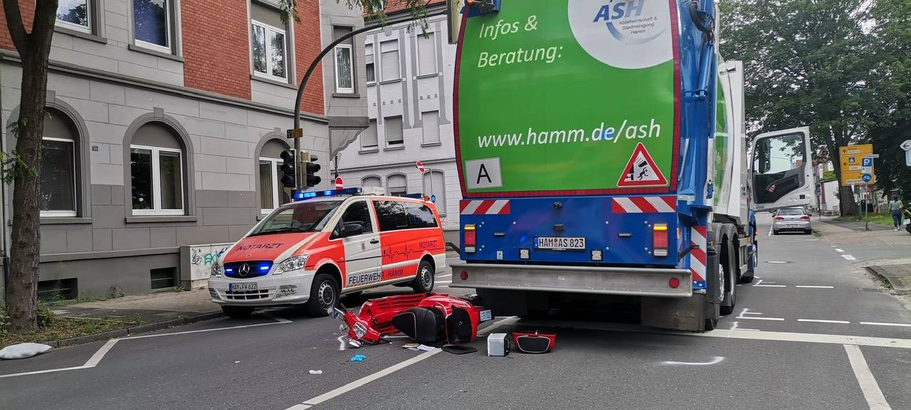 Eine rote Vespa liegt beschädigt auf dem Boden hinter einem Müllwagen. Daneben steht ein Notarztwagen. Der Unfall passierte auf dem Caldenhofer Weg.