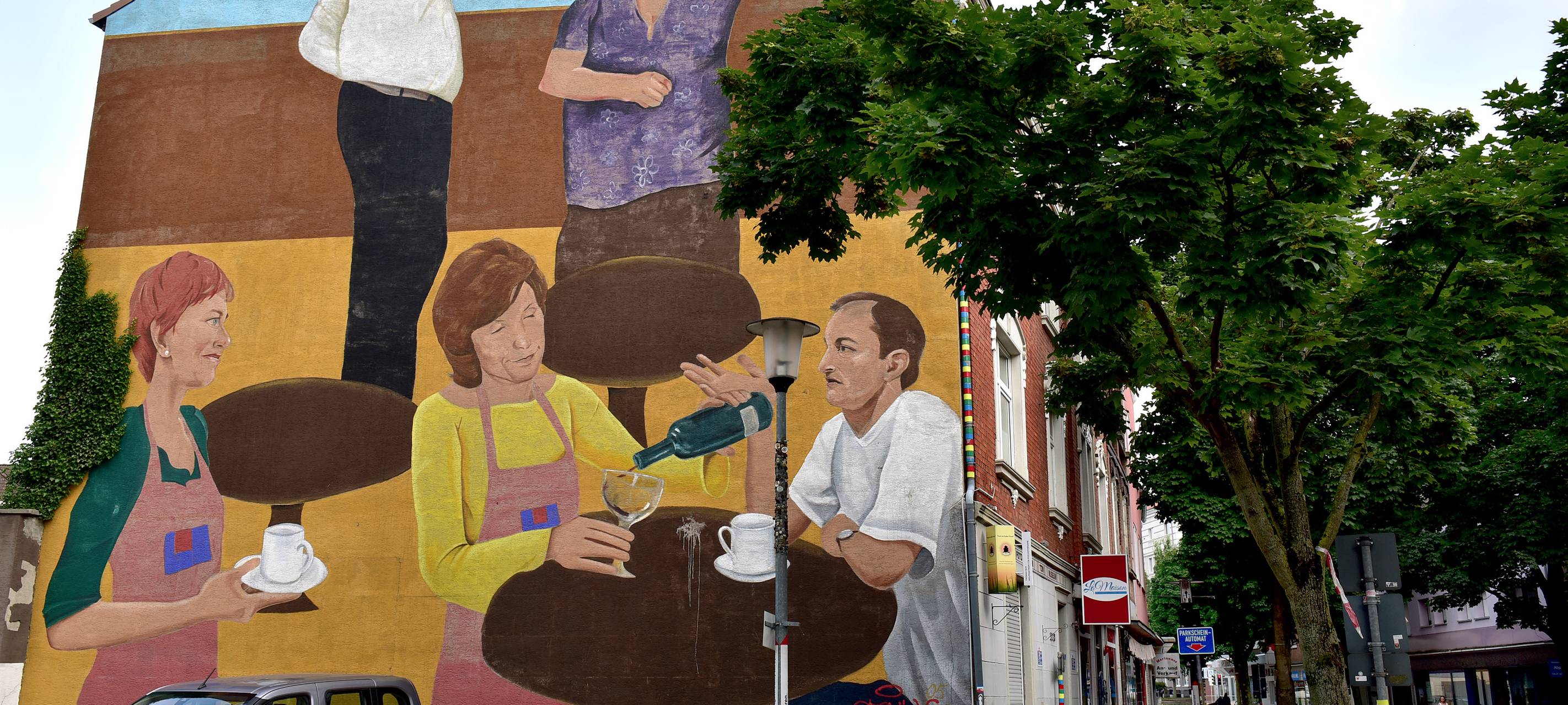 Auf einer großen Hauswandseite im Martin-Luther-Viertel ist ein großes Gemälde. Dort tanzen zwei Menschen, zwei weitere sitzen an einem kleinen Tisch in einem Café.