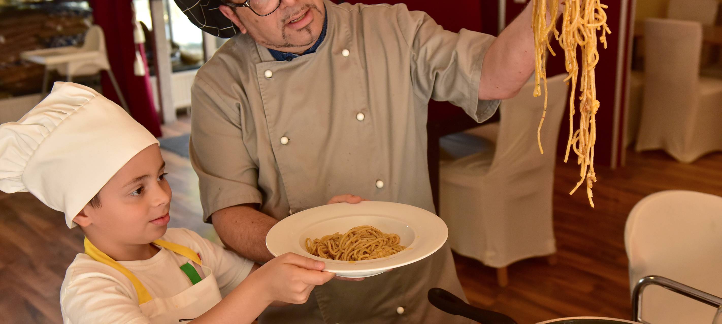 Gastronom Mario hält mit einer Zange eine Portion Spaghetti in der Hand, die er zuvor in einem Käse-Laib gemacht hat. Neben ihm steht ein Kind mit einer weißen Kochmütze, das einen Teller mit ein paar Spaghetti in der Hand hält.