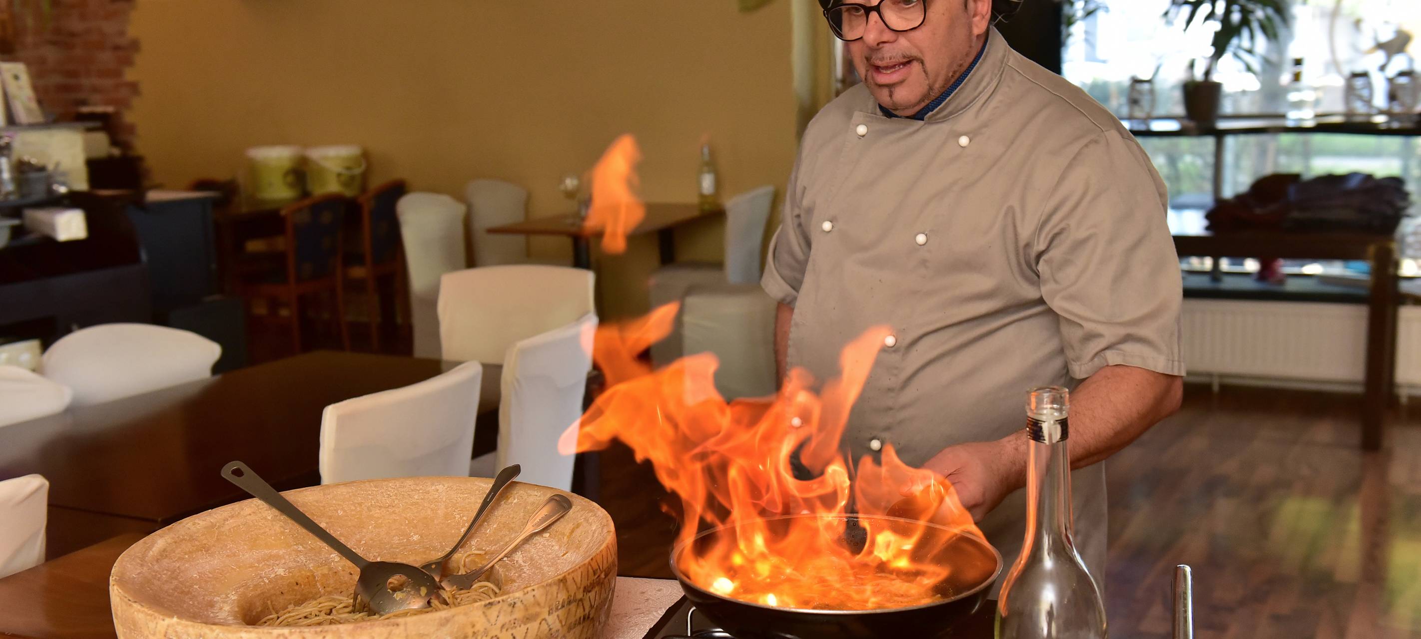 Gastronom Mario steht an einer Pfanne mit Feuer. Neben ihm ist ein großer Laib Käse.