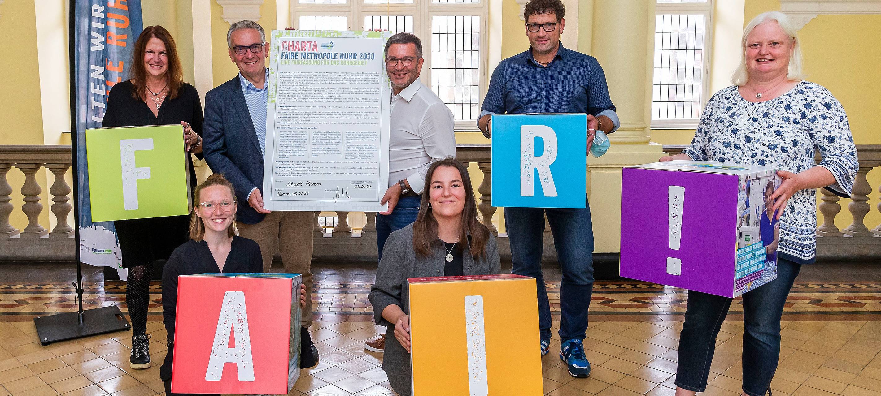 Mehrere Menschen sitzen und stehen im Rathausflur und halten bunte Würfel hoch, auf denen jeweils ein Buchstabe ist. Zusammengesetzt ergebn sie das Wort "Fair".