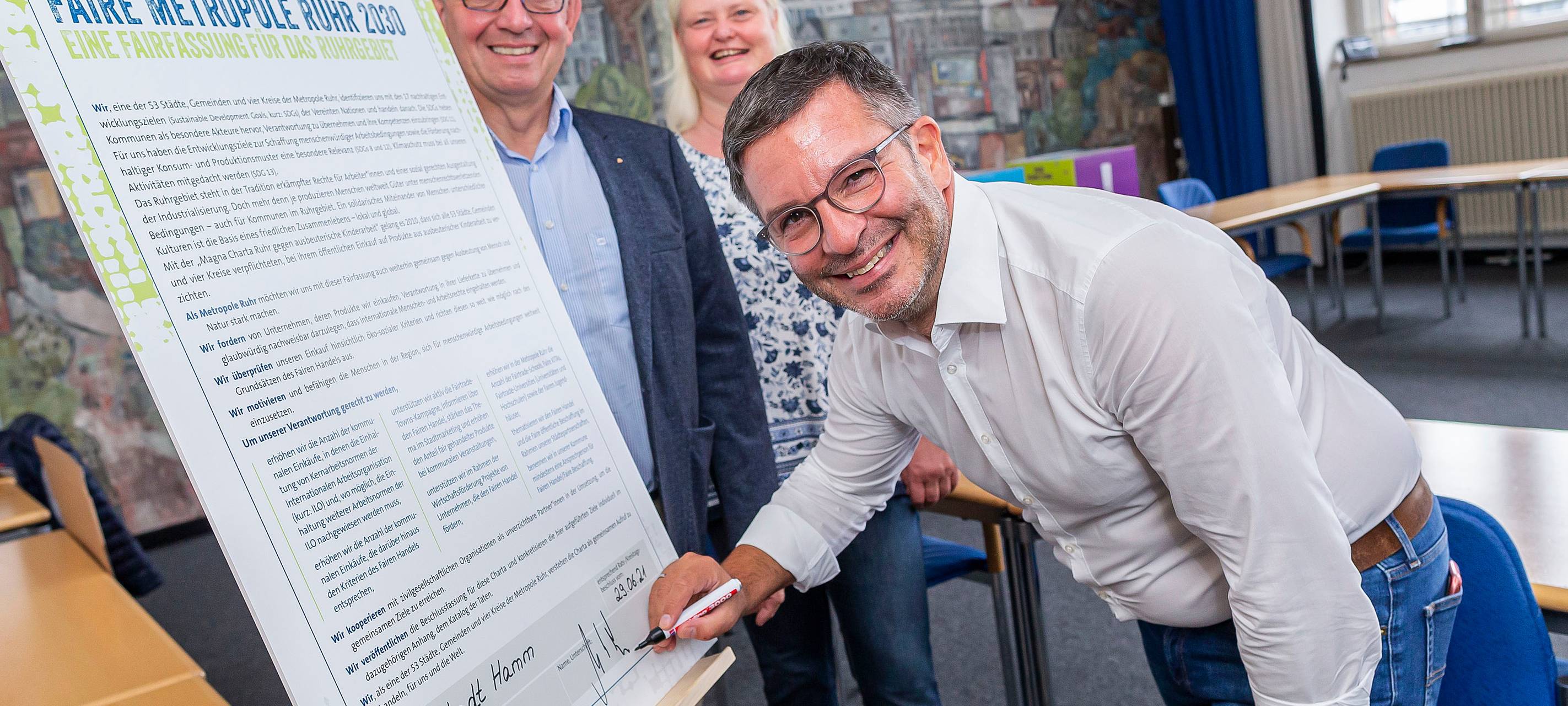 Marc Herter steht vor einer großen Tafel - der Charta Faire Metropole Ruhr. Die unterzeichnet er. Im Hintergrund stehen zwei weitere Personen.