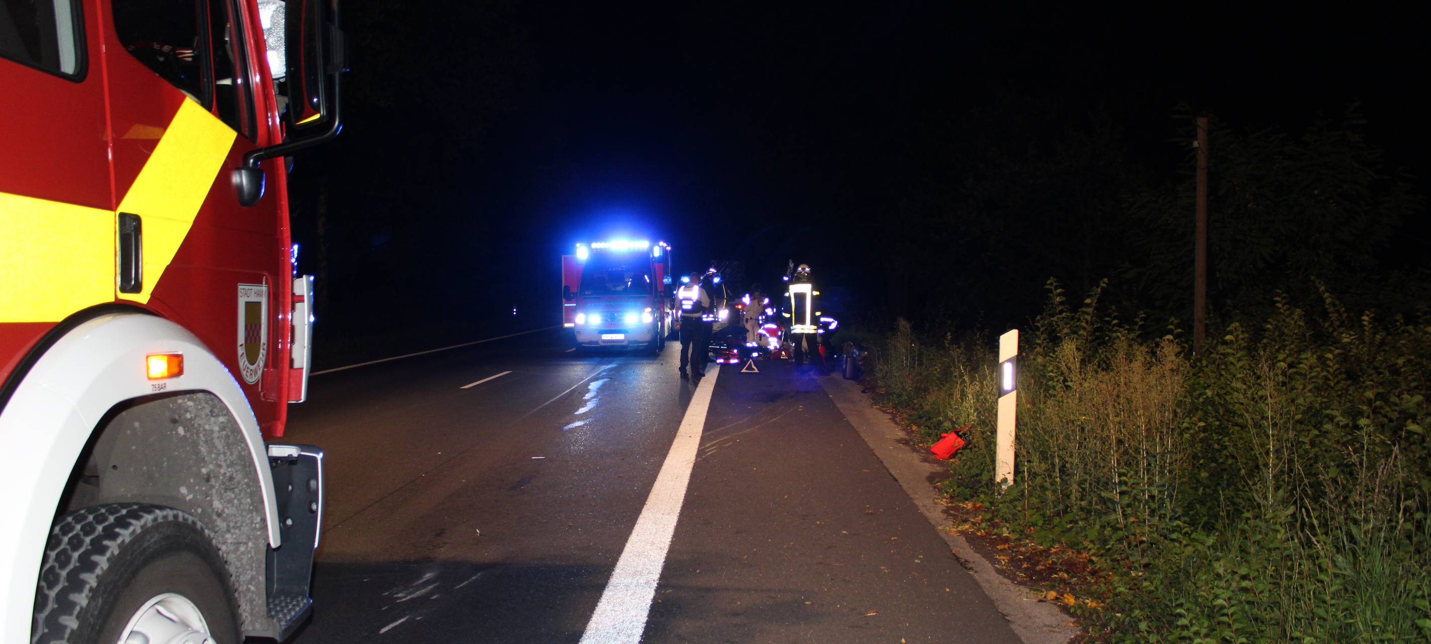 Im Vordergrund ist noch eine Ecke eines Feuerwehrautos. Im hinteren Bereich sieht man mehrere Einsatzkräfte am Boden knien.