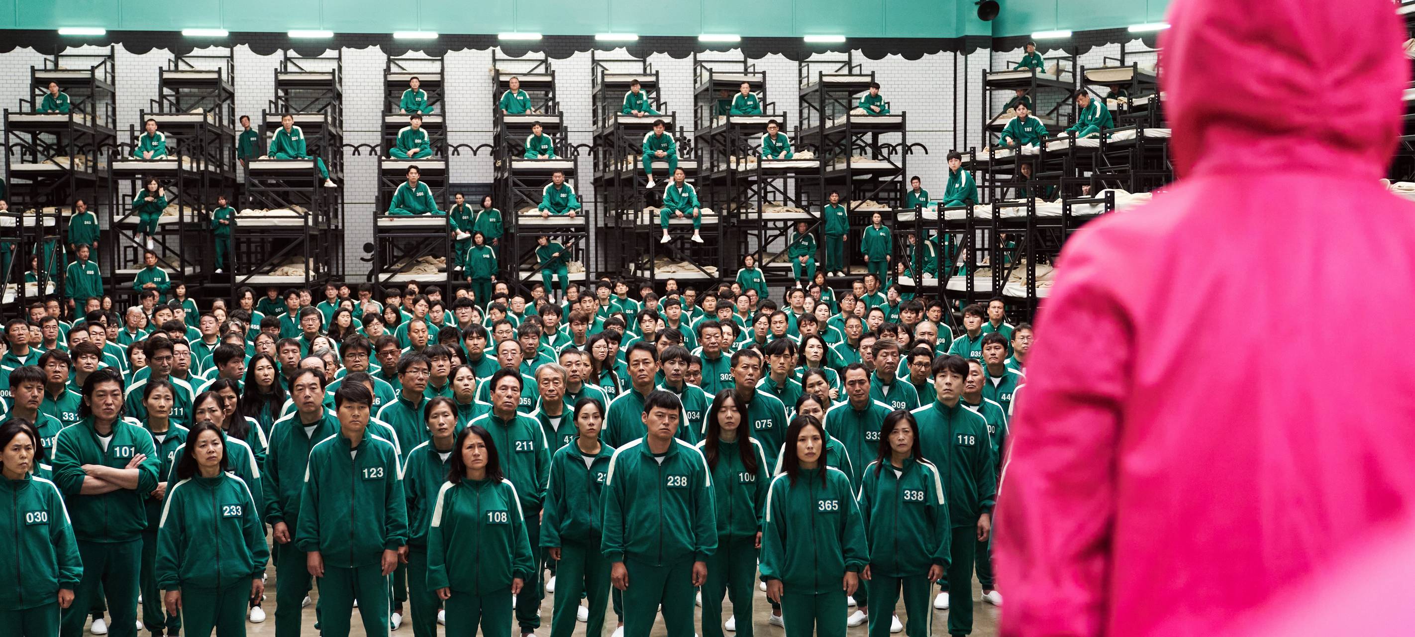 Die 456 Teilnehmer der Serie Squid Game stehen in ihren grün/blauen Anzügen vor einem Mann mit einem roten Anzug.