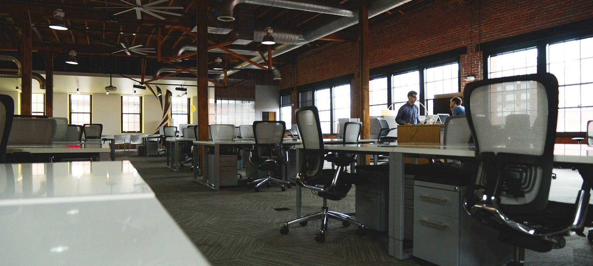 Ein Großraumbüro mit leeren Stühlen und Schreibtischen. Im Hintergrund unterhalten sich nur zwei Menschen.
