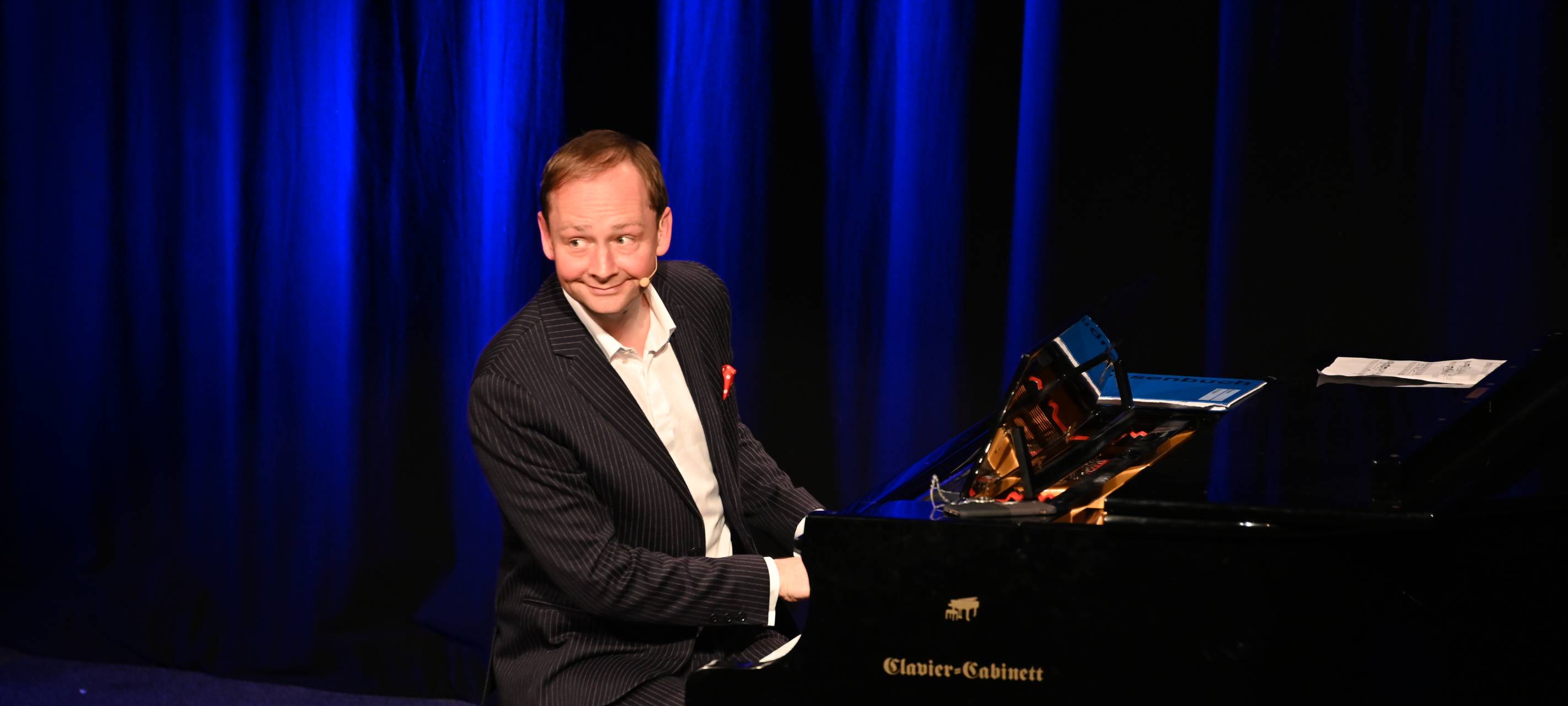 Christoph Reuter sitzt auf einer Bühne am Piano und wendet sich dem Publikum zu