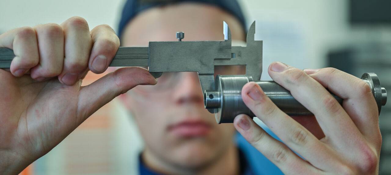 Ein Lehrling zum Mechatroniker prüft mit einem Messschieber ein Werkstück in einer Ausbildungsstätte