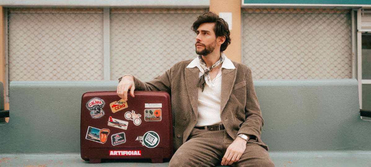 Musiker Alvaro Soler sitzt in einem Pressefoto von 2025 auf einer Bank neben einem Koffer