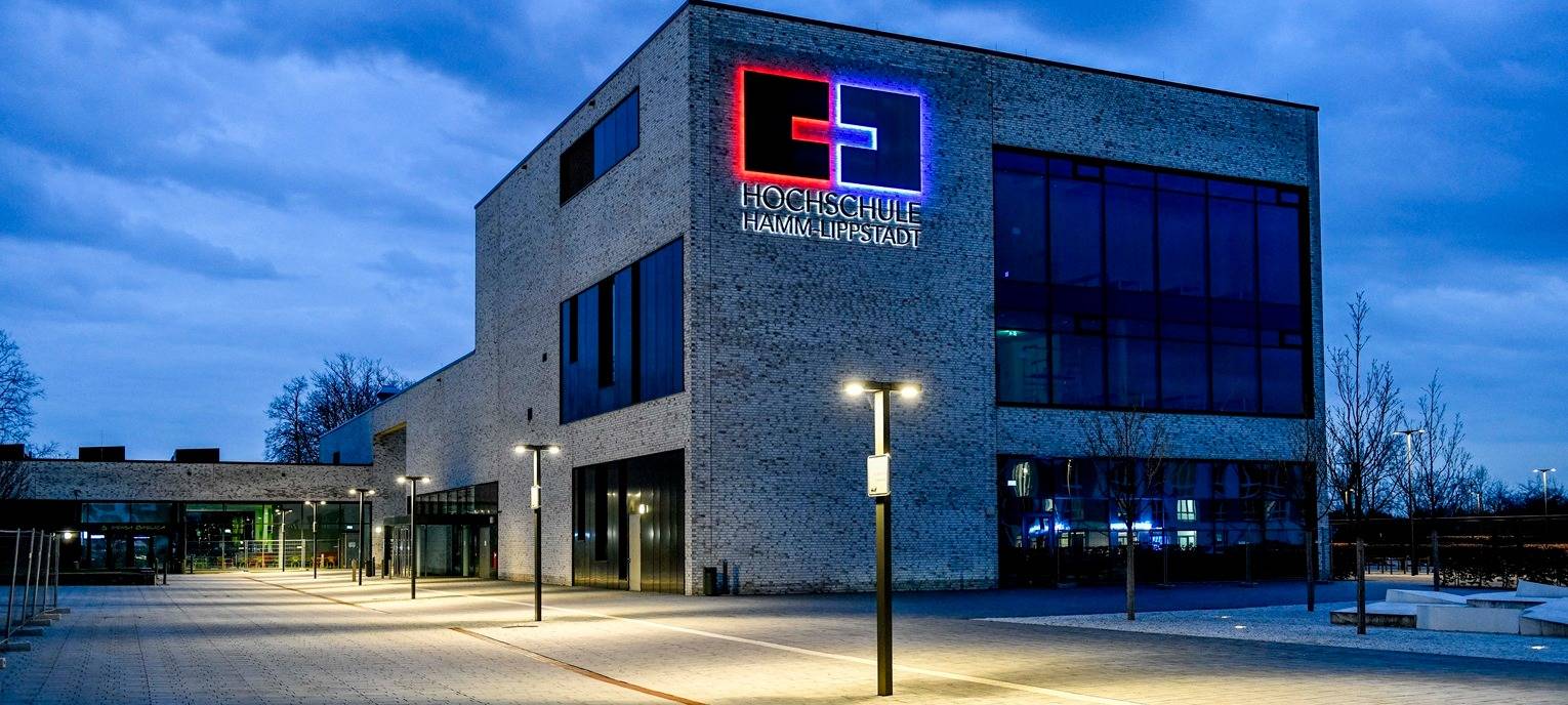 Modernes Hochschulgebäude der HSHL mit beleuchteter Fassade und Logo bei Abenddämmerung, gepflasterter Vorplatz mit Straßenlaternen