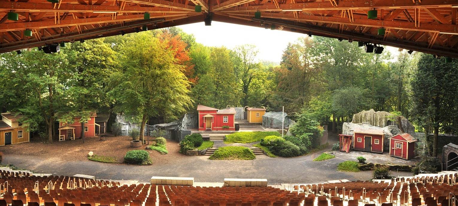Blick auf die Bühne der Waldbühne Heessen mit bunten Bühnenhäusern im Wald, Zuschauerrängen und überdachter Holzkonstruktion