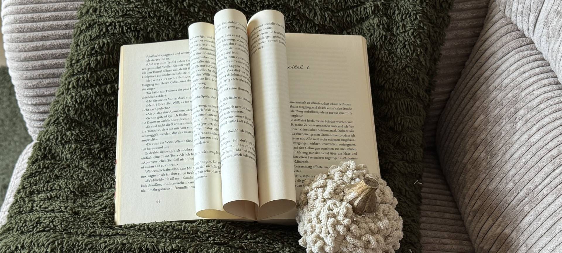 Ein aufgeschlagenes Buch liegt auf einer dunkelgrünen Decke. Daneben ein kleiner gehäkelter Deko-Kürbis