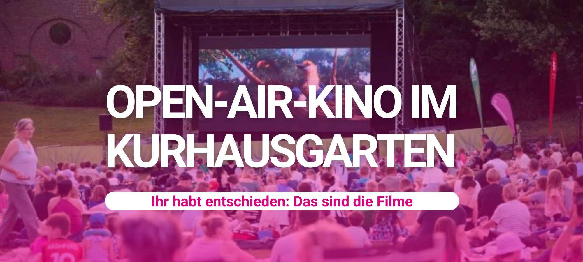 Publikum sitzt beim Open-Air-Kino im Kurhausgarten vor einer beleuchteten Großleinwand, auf der ein Film läuft. Im Vordergrund Besucher auf Decken und Stühlen, im Hintergrund historisches Gebäude und Bäume.